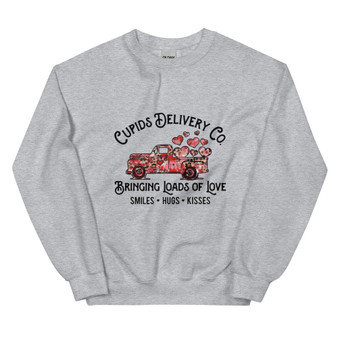 Crew neck sweatshirt in a neutral/pastel color featuring a pickup truck overflowing with red and pink hearts. Text reads “Cupid’s Delivery Co.: Bringing Loads of Love, Smiles, Hugs, and Kisses” in fun, playful lettering. Shown folded and on a flat lay with clear visibility of full design.