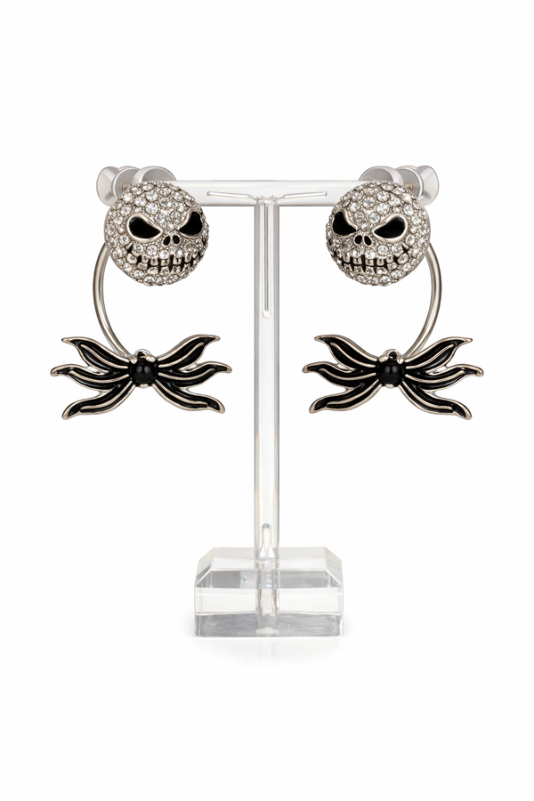 Features skull stud earrings with a separate spider bow backing designed to sit behind the ear, creating a layered, dimensional look. Images include clean white background product shots for detail, a stand display for structure, an on-ear lifestyle photo to show fit and placement, and a dark, moody flat lay to highlight the gothic aesthetic and sparkle.