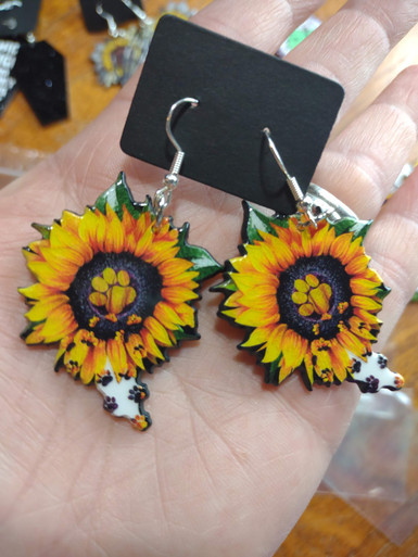 Assorted sunflower-themed acrylic earrings featuring bright yellow petals with dark centers and earthy accents. Each design highlights a rustic, nature-inspired look with bold outlines and clean detailing, displayed on silver fishhook ear wires against a neutral background.