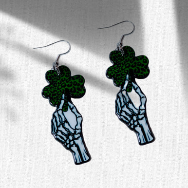 An assorted collection of St. Patrick’s Day-themed acrylic earrings featuring bold, edgy designs with skeletons, skulls, shamrocks, and leprechaun-inspired elements. Each pair showcases vibrant green accents paired with black and white detailing for a high-contrast, gothic-meets-festive aesthetic. Designs include skeleton hands, skull faces, clover details, and playful drinking motifs, all hanging from silver fishhook ear wires. This set blends dark humor, alternative style, and holiday spirit for a unique, statement-making accessory lineup.