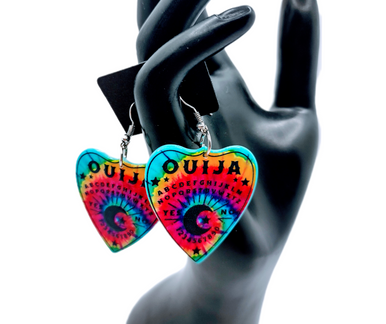 Collection of Ouija planchette acrylic earrings displayed in multiple design variations on a clean background. Each pair features unique lettering styles, symbols, and finishes while maintaining a cohesive dark, witchy aesthetic. The assortment highlights different options within the same occult-inspired theme.