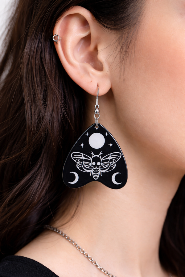 Collection of Ouija planchette acrylic earrings displayed in multiple design variations on a clean background. Each pair features unique lettering styles, symbols, and finishes while maintaining a cohesive dark, witchy aesthetic. The assortment highlights different options within the same occult-inspired theme.