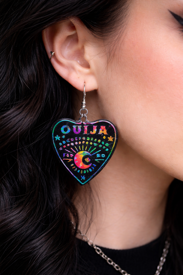 Collection of Ouija planchette acrylic earrings displayed in multiple design variations on a clean background. Each pair features unique lettering styles, symbols, and finishes while maintaining a cohesive dark, witchy aesthetic. The assortment highlights different options within the same occult-inspired theme.