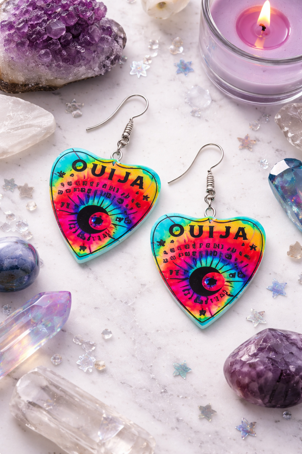 Collection of Ouija planchette acrylic earrings displayed in multiple design variations on a clean background. Each pair features unique lettering styles, symbols, and finishes while maintaining a cohesive dark, witchy aesthetic. The assortment highlights different options within the same occult-inspired theme.