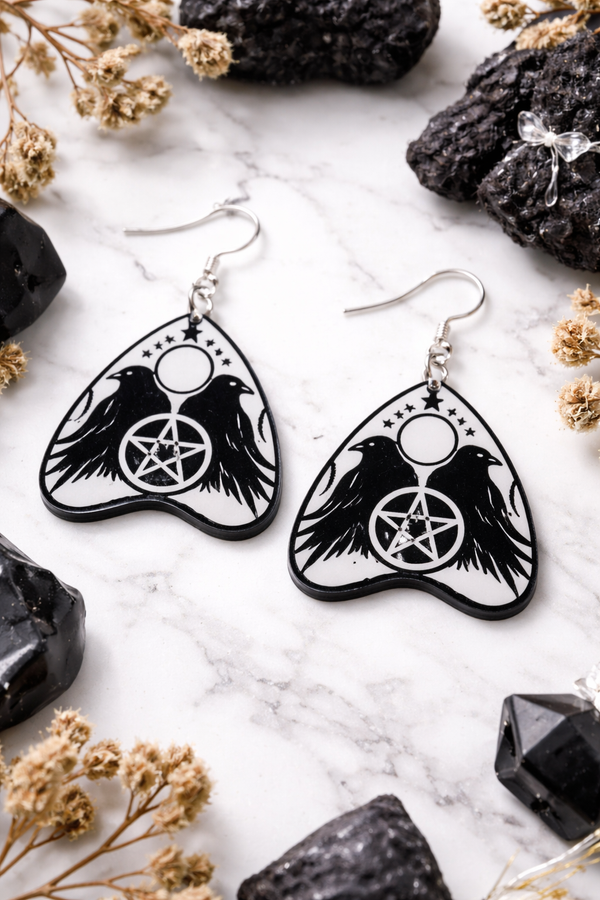 Collection of Ouija planchette acrylic earrings displayed in multiple design variations on a clean background. Each pair features unique lettering styles, symbols, and finishes while maintaining a cohesive dark, witchy aesthetic. The assortment highlights different options within the same occult-inspired theme.
