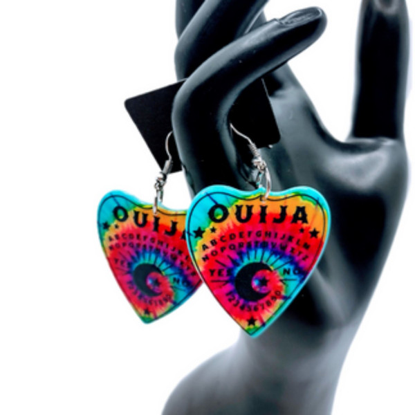 Collection of Ouija planchette acrylic earrings displayed in multiple design variations on a clean background. Each pair features unique lettering styles, symbols, and finishes while maintaining a cohesive dark, witchy aesthetic. The assortment highlights different options within the same occult-inspired theme.