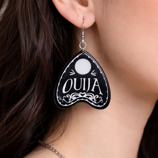 Collection of Ouija planchette acrylic earrings displayed in multiple design variations on a clean background. Each pair features unique lettering styles, symbols, and finishes while maintaining a cohesive dark, witchy aesthetic. The assortment highlights different options within the same occult-inspired theme.