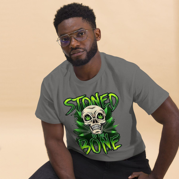 Charcoal gray unisex tee featuring a neon green “Stoned to the Bone” skull design with cannabis leaf eyes and grunge-style lettering, centered on the chest.