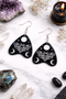 Collection of Ouija planchette acrylic earrings displayed in multiple design variations on a clean background. Each pair features unique lettering styles, symbols, and finishes while maintaining a cohesive dark, witchy aesthetic. The assortment highlights different options within the same occult-inspired theme.