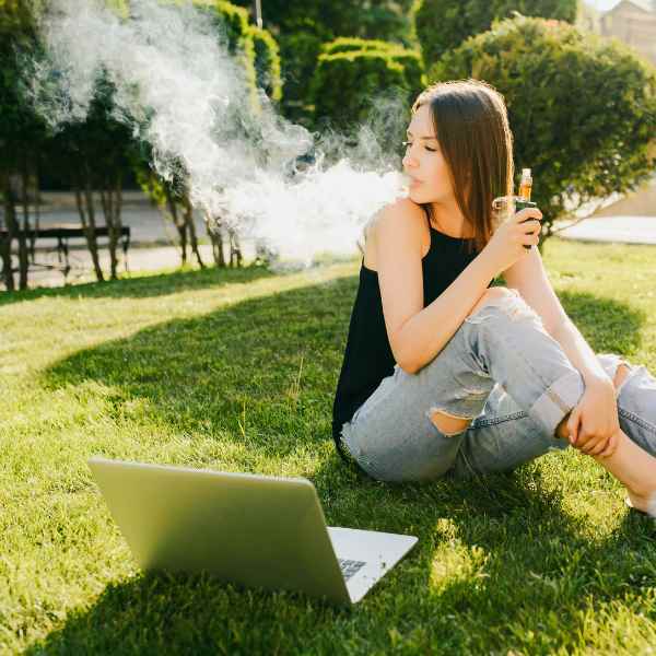 YOung woman vaping while sat on grass in front of a laptop