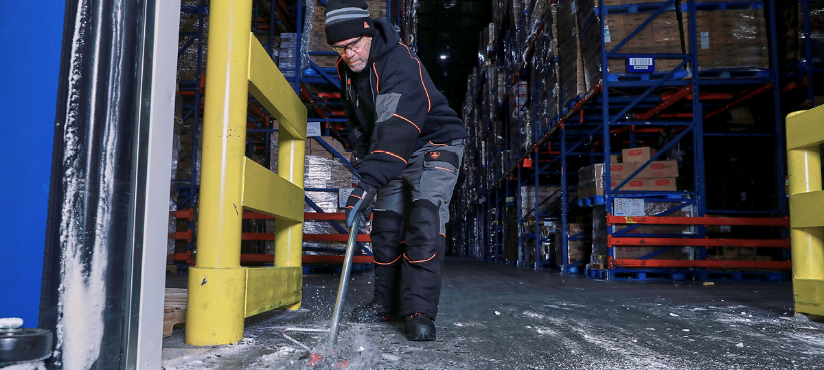 Employee working in frozen cold storage warehouse facility wearing insulated workwear while scraping ice off floors.