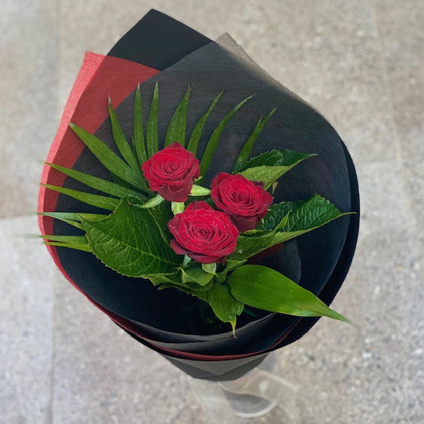 Three red roses and seasonal foliage, wrapped in black paper.