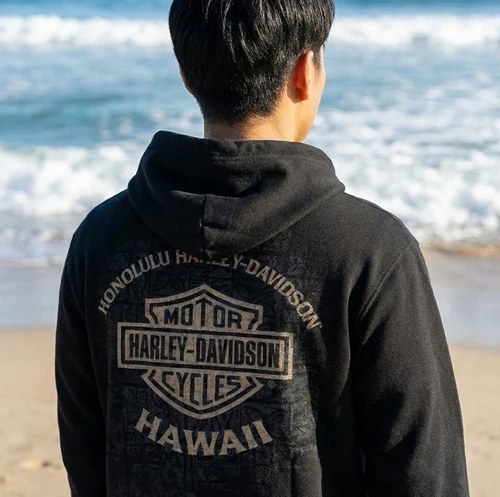 Back view of black Harley-Davidson Men's Tapa Shield Zip-Up Hoodie showing large Honolulu Harley-Davidson Hawaii bar and shield back graphic against ocean backdrop