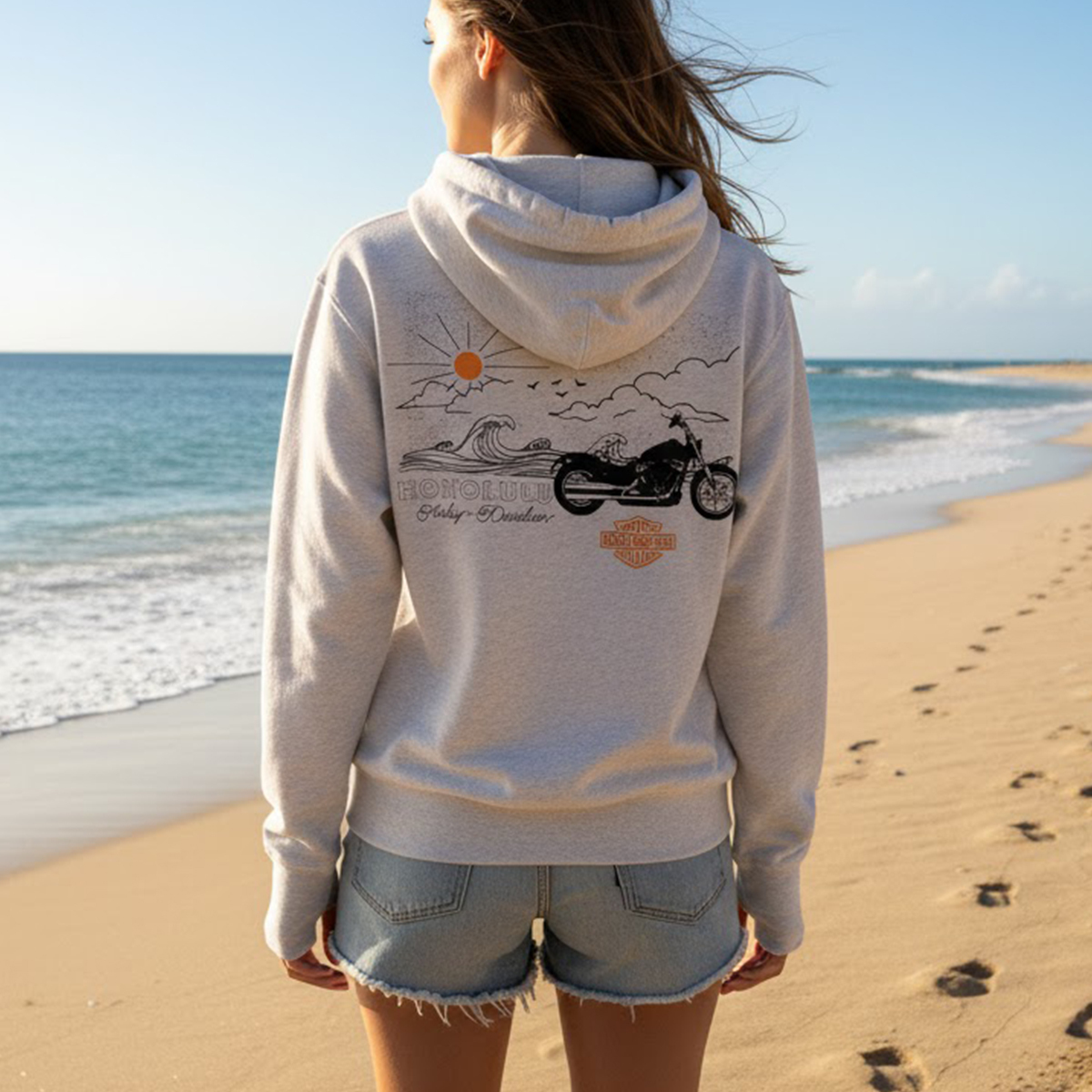 Women's Harley-Davidson Coastal Blue Aloha Blouse Top, a sleeveless blue tropical print top, modeled on a Hawaiian beach with a yellow hibiscus flower.