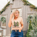 Oh Christmas Tree Oh Christmas Tree DTF Heat Transfer, a woman wearing a light-colored t-shirt with a Christmas tree design and the text 'Oh Christmas Tree', standing outdoors near a festive wooden house.