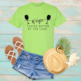 Wine Tastes Better At The Lake DTF Heat Transfer, a lime green T-shirt with wine glass graphics and text, summer fashion flat lay with denim shorts, sandals, a straw hat, seashell, and tropical leaves on a rustic wooden background.