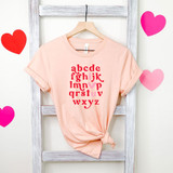 Vday Alphabet DTF Heat Transfer, pink t-shirt with red and pink alphabet letters and a pink heart in the middle, displayed on a hanger against a white wooden background with pink and red heart decorations.