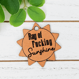 Ray of Fucking Sunshine Tan Leather Keychain with sunburst design and black lettering on a white wooden surface with green leaves