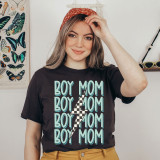Boy Lightening Bolt Mom DTF Heat Transfer, woman wearing a black T-shirt with a multiple blue 'Boy Mom' text and lightning bolt design, indoor setting with butterfly poster and sunglasses in background.