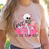 Love Is In The Air Skellie DTF Heat Transfer, a graphic of a skeleton wearing a gas mask and surrounded by pink hearts and splashes on a pink T-shirt.