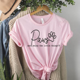 Paws And Enjoy The Little Things DTF Heat Transfer, pink T-shirt with dog paw print and text design, displayed hanging on a wooden hanger with clothing and greenery around.
