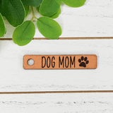 Dog Mom Leather Keychain, a brown leather keychain engraved with 'Dog Mom' and a paw print, placed on a white wooden surface next to green leaves.