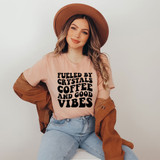 Fueled By Crystals Coffee And Good Vibes DTF Heat Transfer, a woman wearing a pink T-shirt with the text printed in bold black letters, a brown hat, and a brown jacket, smiling and sitting against a plain white background.