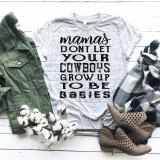 Mamas Don't Let Your Cowboys Grow Up To Be Babies Sublimation Transfer, a printed T-shirt with text about mothers and cowboys, surrounded by fashion items like an olive green jacket, plaid scarf, tan shoes, and cotton branches on a white wooden background.