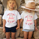 Don't Worry Be Hoppy Pink DTF Heat Transfer, two young girls wearing white T-shirts with bunny graphic and pink sunglasses design, standing outdoors against a stone wall.
