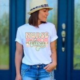 Nurse Vibes Retro DTF Heat Transfer, a woman wearing a white t-shirt with colorful Nurse Vibes design and a beige hat with a floral band, standing outdoors in front of blue doors.