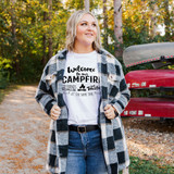 Welcome To Our Campfire DTF Heat Transfer, a woman outdoors wearing a white T-shirt with black campfire themed design and inspirational text, surrounded by autumn trees and canoes.