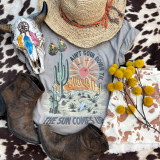 Ain't Going Down Screen Print Heat Transfer, colorful desert-themed graphic with cactus and sun elements on a gray t-shirt, complemented by cowboy boots, a straw hat, turquoise earrings, a painted skull, yellow flowers, and cowhide fabric.