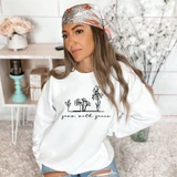 Grow With Grace DTF Heat Transfer, woman wearing a white sweatshirt with black floral line art and text, in a cozy decorated interior setting.