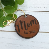 Custom Mom Leather Keychain, personalized round brown leather keychain with 'Mom' text and 'Est. 2021' engraving, attached to a gold key ring and clasp, on a white wooden surface with green leaves.