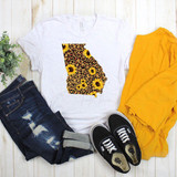 Georgia Leopard and Sunflower Sublimation Transfer, white t-shirt with a leopard print and sunflower design, paired with distressed jeans, yellow jacket, and black Vans sneakers on a wooden background.