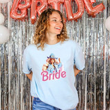 Bride Barbie DTF Heat Transfer, a woman wearing a light blue T-shirt with a Barbie bride graphic and the word 'Bride' printed on it, standing in front of silver tinsel background with pink balloon letters spelling 'BRIDE'.