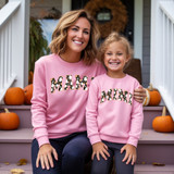Mama Floral Ghost DTF Heat Transfer, image of a smiling woman and young girl wearing matching pink sweatshirts with Halloween-themed 'MAMA' and 'MINI' designs, sitting on steps decorated with pumpkins for fall.