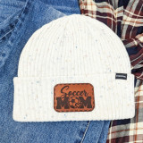 Soccer Mom Leather Hat Patch, A brown leather patch with the words 'Soccer Mom' and a soccer ball design, sewn onto a white ribbed knit beanie.