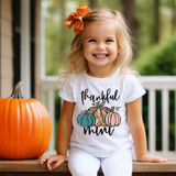 Thankful Mini DTF Heat Transfer, a smiling young girl wearing a white t-shirt with colorful pumpkins and the words 'thankful mommy', sitting outdoors next to a large pumpkin.