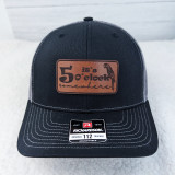 It's 5 o'clock Somewhere Parrot Leather Hat Patch, a stylish trucker cap with a leather patch embroidered with the phrase 'It's 5 o'clock somewhere' and a parrot illustration, featuring a black crown and brim with gray mesh sides.