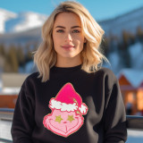 Pink Grouchy Guy Chenille Patch, eye-catching pink patch with a grumpy face wearing a Christmas hat on black sweatshirt worn by a blonde woman outdoors in a snowy mountain setting.