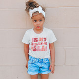 In My Nutcracker Era Sublimation Transfer, a young girl wearing a white t-shirt with pink text and nutcracker graphics stands against a beige wall.
