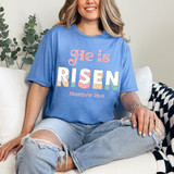 He Is Risen Matthew 28:6 DTF Heat Transfer, colorful and vibrant Bible verse design on a blue T-shirt worn by a woman sitting on a couch.