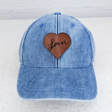 Lover Leather Hat Patch, blue denim baseball cap with a brown leather heart patch and the word 'lover' embroidered in black cursive on the patch.