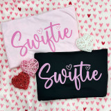 Swiftie DTF Heat Transfer, pink and black t-shirts with 'Swiftie' design and heart accents, on pink heart-patterned fabric with woven heart decorations.