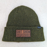 Shit Show Supervisor Leather Hat Patch, olive green knit beanie with a leather patch saying 'SHIT SHOW SUPERVISOR' and a small black tag with white text 'PACIFIC'.