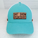 Shit Show Supervisor Leather Hat Patch, turquoise trucker cap with pink and brown detailing, and a beige patch with black text that reads 'Shit Show Supervisor'.
