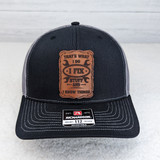 That's What I Do Leather Hat Patch, close-up of a black and gray trucker hat with a brown leather patch featuring the quote 'That's What I Do I Fix Stuff And I Know Things' in black text.