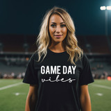 Game Day Vibes WHITE DTF Heat Transfer, a woman wearing a black t-shirt with 'GAME DAY vibes' text printed on it, standing on a sports field at night.