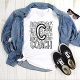 Coach Typography Sublimation Transfer, a white t-shirt with black hand-drawn style coach letter and logo design, laid flat with a denim jacket, black sunglasses, and black Vans sneakers on a wooden surface.